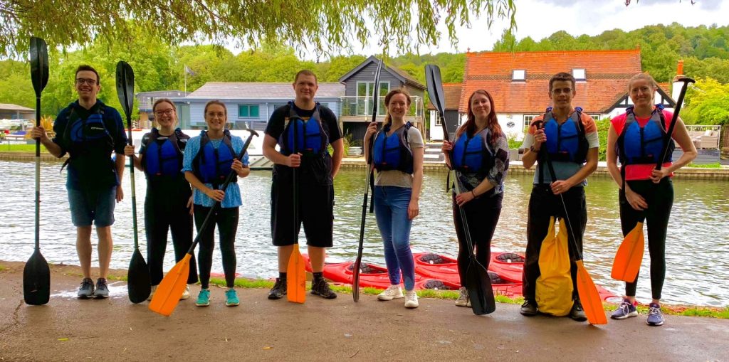 A group of young adults standing by the river holding their canoe paddles and wearing blue life vests